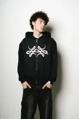 Hoodie con cierre frontal, color negro, de corte regular fit. Presenta un estampado frontal grande en blanco con un diseño abstracto y estilizado, similar a un monograma o símbolo.