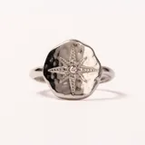 Anillo plateado con diseño de estrella de ocho puntas en el centro, con un pequeño cristal en el medio. El anillo tiene una superficie texturizada y un acabado brillante.