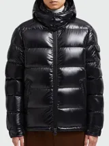 Campera corta de plumas Moncler modelo Maya, color negro con acabado brillante. Presenta diseño acolchado, capucha integrada, cierre frontal con cremallera y bolsillo en la manga.