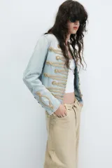 Chaqueta de mezclilla clara estilo militar, con cuello redondo, manga larga y detalles de cordones y botones dorados en el frente.