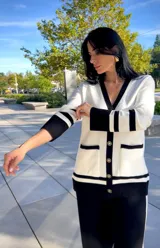 Cardigan de punto en color blanco con detalles en negro en los bordes, cuello en V, cierre frontal con botones dorados y bolsillos delanteros.