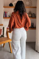 Camisa de color terracota con cuello camisero, cierre frontal de botones y mangas largas abullonadas. Presenta un delicado bordado floral en color blanco en la zona del pecho y los hombros, con un diseño de corte holgado.