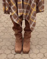 Botas de caña alta confeccionadas en cuero gamuzado color marrón. Presentan un diseño de estilo western con puntera cuadrada, taco de madera y una tira decorativa con estribo metálico desmontable en la zona del tobillo.