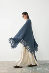 Ruana triangular tejida en lana merino extrafina, lino y viscosa, con textura aireada y terminación de flecos en los bordes. Diseño versátil y envolvente.