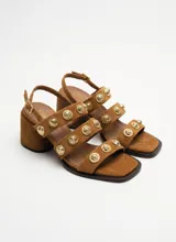 Sandalia de cuero gamuzado color marrón, con tres tiras delanteras decoradas con apliques metálicos dorados en relieve, taco cuadrado y tira ajustable en el talón con hebilla dorada.