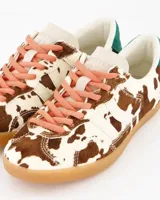 Sneakers de estilo urbano con estampado animal print tipo vaca, confeccionadas en material con textura de pelo. Presentan cordones en tono rosado, detalle de talón en color verde y suela de goma color caramelo.