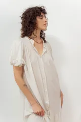Vestido midi color crudo, con cierre frontal de botones, escote en V y mangas cortas abullonadas. Presenta un diseño holgado con detalle de volados en el bajo.