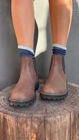 Bota tipo Chelsea de cuero marrón con textura granulada, paneles laterales elásticos en color negro y suela track dentada con costura blanca visible.