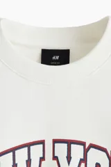 Buzo de algodón con cuello redondo, hombros caídos y puños acanalados. Presenta un estampado frontal con estilo universitario que incluye el texto 'NEW YORK PHYS.ED.' y un emblema central en tonos rojo y azul.