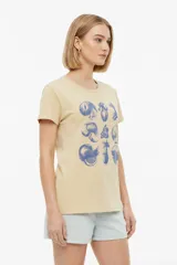 Remera blanca de manga corta y cuello redondo, con diseño estampado en el frente que simula un sello postal con palmeras y la inscripción 'la paloma'.