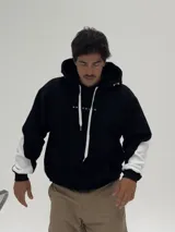 Canguro con capucha de corte boxy, color negro con una franja horizontal blanca que recorre la espalda y las mangas, presentando la palabra 'BROOKLYN' estampada en negro sobre la franja. La capucha también incluye detalles estampados.