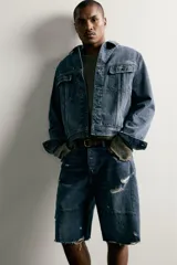 Bermuda baggy fit confeccionada en denim de algodón con lavado azul y efecto rotos en toda la prenda. Presenta diseño de cinco bolsillos, detalles de costuras en las perneras, bajo acabado irregular y cierre frontal con cremallera y botón.