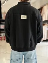 Buzo gris oscuro de corte oversize con cuello alto y cierre frontal parcial. Cuenta con mangas largas y puños elásticos, además de un pequeño bordado con texto en el pecho.