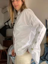 Camisa blanca de popelina de algodón, de corte oversize, con cuello camisero, manga larga y detalles de botones decorativos en las mangas.