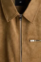 Campera de gamuza sintética color tostado, con cuello camisero, cierre frontal, bolsillo superior con cierre y bolsillos laterales ribeteados.