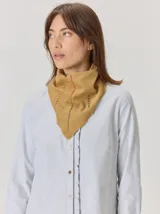Bandana de forma triangular en tono gris oscuro con estampado de cuadros sutiles. Presenta detalles de pespuntes a mano en hilo naranja y blanco, junto con pequeños apliques decorativos que le otorgan un estilo artesanal y único.