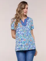 Blusa de manga corta con escote en V y pechera bordada. Presenta un estampado multicolor sobre base blanca, con pliegues frontales y aberturas laterales en el ruedo.