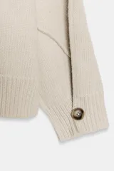 Jersey de punto color beige con cuello subido, manga larga y detalle de botones en los puños. Presenta costuras marcadas y acabados en rib.