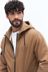 Campera de felpa color camel con cierre frontal, capucha ajustable con cordones negros y bolsillos laterales con cierre negro. Presenta un estampado pequeño en el pecho con texto en color oscuro.
