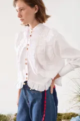 Camisa de lino blanca con cuello mao, manga larga y botones frontales. Presenta volados delanteros con terminaciones desflecadas y detalles de costuras en hilo rojo.