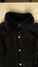 Camisa de cuero negro con cuello de corderito natural, cierre frontal con broches metálicos y puños ajustables.