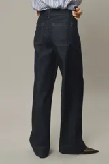 Pantalón de jean de denim oscuro, tiro alto, con pierna ancha y corte recto desde la cadera hasta el bajo. Presenta pliegues frontales y traseros, y un dobladillo ancho en los tobillos. Cierre frontal con dos botones dorados.