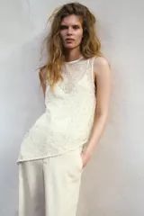 Top sin mangas de cuello redondo, confeccionado en tejido cubierto de lentejuelas brillantes. Presenta un diseño asimétrico en el bajo con una caída lateral pronunciada y cuenta con un forro interior con tirantes finos. Incluye cierre en la espalda con abertura y botón oculto.