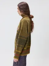 Pullover de textura tipo terciopelo con cuello alto y cierre frontal parcial. Presenta un diseño a rayas horizontales en tonos grises y negros, con detalles en rojo y blanco en el cuello, puños y ruedo. Incluye un aplique bordado con la letra M en el pecho y puños acanalados.