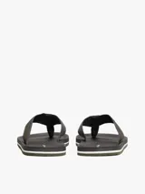 Ojotas para hombre con base oscura, suela de EVA y tiras de textil trenzado en tonos gris y verde oliva. Presentan un parche con el logo de Tommy Hilfiger en la tira y líneas decorativas blancas y verdes en el borde de la suela.