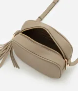 Cartera bandolera pequeña color beige, confeccionada en poliuretano con textura granulada. Presenta cierre superior con cremallera, correa larga ajustable y detalle de borla decorativa en el lateral con herrajes dorados.