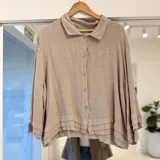 Camisa de lino color beige, de corte holgado, con cuello camisero, cierre frontal de botones y detalles de volados horizontales en el ruedo y los puños de las mangas largas.