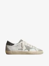 Championes Golden Goose modelo Super-Star, confeccionados en cuero color blanco con paneles de gamuza gris. Presentan el icónico parche de estrella lateral, detalle de glitter en el talón, efecto envejecido en la suela y logo GGDB grabado en el lateral.
