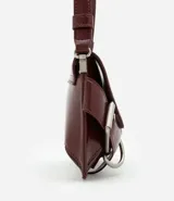 Cartera tipo baguette de tamaño pequeño, confeccionada en poliuretano color negro. Presenta una solapa frontal con detalles de hebillas metálicas decorativas en los laterales y un asa de hombro ajustable.