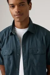 Camisa de manga corta color azul petróleo, con cuello clásico, cierre frontal con botones, dos bolsillos de parche con botón en el pecho y corte recto.