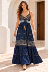 Vestido largo de denim azul oscuro, estilo solera con tirantes finos, escote en V y cordones con borlas en el centro del pecho. Presenta detalles bordados en hilo dorado en el busto y en la parte inferior de la falda, con diseño en niveles y volados.