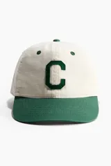 Gorra de sarga de algodón de visera curva, color beige con visera y detalles en verde. Presenta una letra C bordada en el frente, ojales bordados y botón superior en contraste.