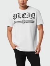 Camiseta blanca de manga corta con cuello redondo. Estampado frontal con la palabra "PLEIN" en tipografía gótica negra, flanqueada por dos líneas horizontales y un emblema central con detalles florales y texto pequeño.