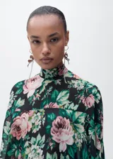 Blusa de gasa con estampado floral en tonos rosa, verde y negro. Tiene cuello alto, mangas largas con volantes y cierre de lazo en la parte posterior.