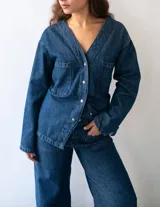 Camisa de denim de algodón con cuello en V, cierre frontal con botones metálicos y bolsillos cuadrados en el pecho. Presenta un diseño con ajuste en la espalda que permite entallar la silueta.