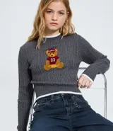 Sweater cropped de punto gris con diseño trenzado, cuello redondo, puños y ruedo con ribetes blancos. Presenta un aplique bordado en el pecho con diseño de osito deportista.