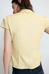 Top de cuello solapa y escote en pico, con manga corta y cierre frontal con botones. Presenta un diseño ajustado con detalles de frunces en el delantero.