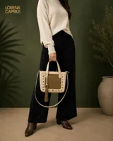 Bolso de mano tipo tote pequeño, confeccionado en cuero vacuno con textura crocco en color arena y panel central en gamuza verde oliva. Presenta detalles de tachas metálicas plateadas en el frente, borla decorativa con argolla y correa larga desmontable y ajustable.