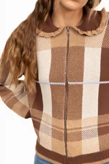 Campera de punto con diseño a cuadros tartán en tonos marrones y beige. Presenta un corte oversize, cierre frontal con cremallera, cuello tipo babero con volados y mangas largas con puños ajustados.