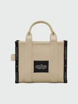 Cartera tipo tote bag de lona color beige, con diseño estructurado y asas superiores. Presenta el logo de la marca estampado en la parte frontal y detalles de cintas laterales con inscripciones.