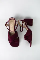 Sandalia de tacón cuadrado medio, confeccionada en material textil tipo gamuza color negro. Presenta un diseño de tiras cruzadas con nudo en la parte delantera y una tira ajustable con hebilla metálica en el tobillo.