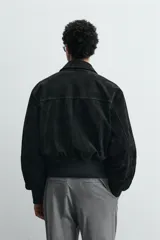 Cazadora cropped fit de piel con acabado serraje y textura de ante, color negro. Presenta cuello camisero, cierre frontal con cremallera, bolsillos delanteros de plastrón con solapa en el pecho y acabado de rib elástico en los puños y el bajo.