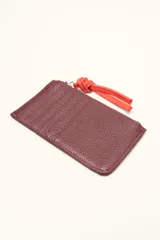 Monedero rectangular de cuero vacuno con textura granulada en color marrón topo. Cuenta con cierre superior metálico y un tirador decorativo de cuero en color naranja con un nudo. Presenta un pequeño logo grabado en el centro.