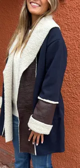 Saco de paño azul marino con solapas amplias y puños en corderito sintético color crema. Presenta detalles en cuero sintético marrón en la parte frontal y en las mangas, con un diseño abierto y corte recto.