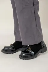 Bota corta de cuero negro con suela track, cierre frontal con cremallera y tira decorativa con hebilla metálica sobre el empeine.