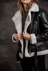 Campera tipo gamulán de eco cuero negro con interior y solapas de corderito sintético en color crudo. Presenta cierre frontal, bolsillos laterales con cierre y cinturón ajustable en la cintura.